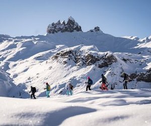 People with skiing equipment are hiking through a snowy mountain landscape under a clear sky.