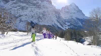 People in colorful winter clothing are hiking in the snow in front of a mountain backdrop.