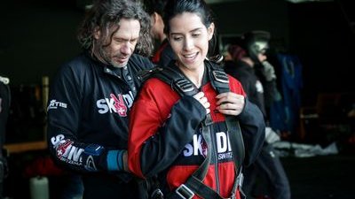 Personen mit Fallschirmgurten in einem Innenraum, eine Person hilft der anderen beim Anlegen der Ausrüstung.