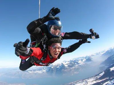 Two people tandem skydiving over snow-covered mountains, wearing helmets and goggles.