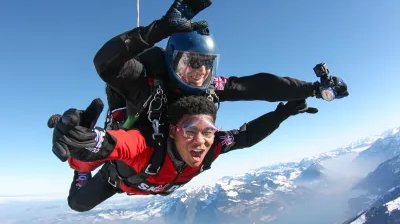 Two people tandem skydiving over snow-covered mountains, wearing helmets and goggles.