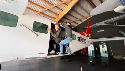 Two people board an airplane in a hangar, visible are airplane doors and wings.