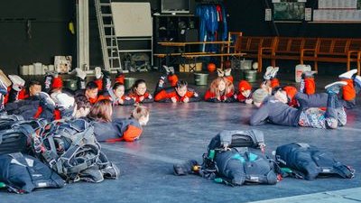 Personen in Overalls liegen auf dem Boden in einer Halle, umgeben von Fallschirmen und Ausrüstung.