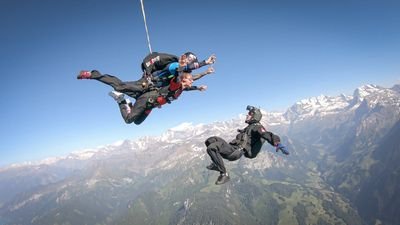 Skydiver in the air with helmet and suit over mountain landscape, accompanied by a guide.