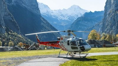 Ein roter Helikopter steht auf einer Wiese in einem alpinen Tal mit hohen Bergen im Hintergrund.
