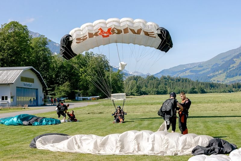 Parachutists land on a meadow in front of a hangar in the Swiss Alps, with parachutes.