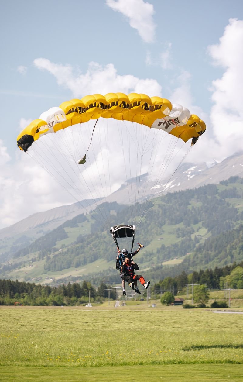 Two people with parachutes land on a meadow surrounded by mountains.