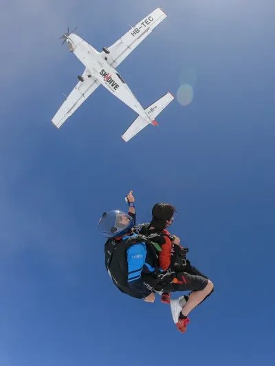 Two people in a tandem skydiving jump with helmets and harnesses, airplane in the background, blue sky.