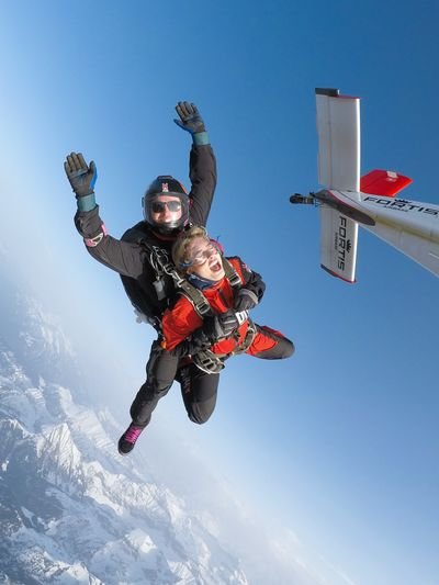Tandem skydivers in freefall over snowy mountains, wearing helmets and harnesses, near an airplane.