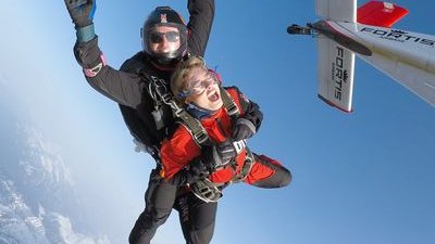 Tandem-Fallschirmspringer im freien Fall über schneebedeckten Bergen, tragen Helme und Gurte, in der Nähe eines Flugzeugs.