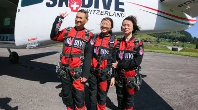 Three people in skydiving suits are standing next to an airplane on an airfield.