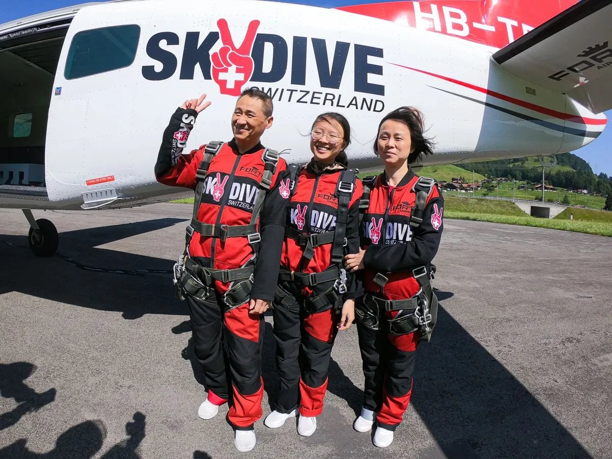 Drei Personen in Fallschirmsprunganzügen stehen neben einem Flugzeug auf einem Flugplatz.
