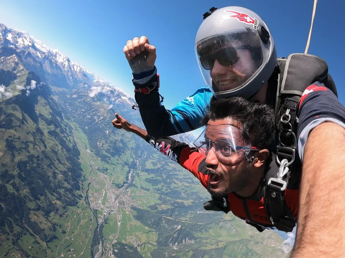 Zwei Personen beim Tandem-Fallschirmsprung über einer Berglandschaft, mit Helmen und Schutzbrillen.