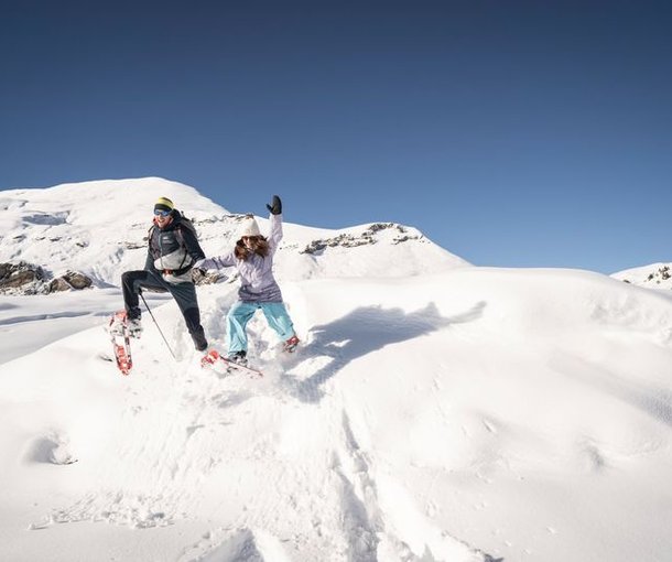 Two people in snowshoes jumping on a snowy mountain slope under a clear blue sky.