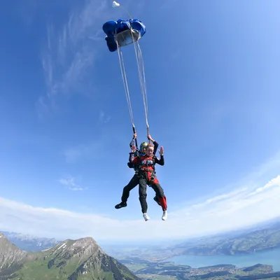 Two people tandem skydiving over mountains, with blue parachutes and safety equipment.