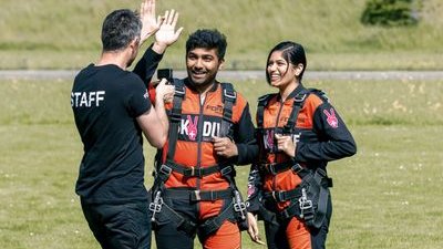Zwei Personen in Fallschirmspringeranzügen mit Gurten auf einer Wiese, begleitet von einem Mitarbeiter.